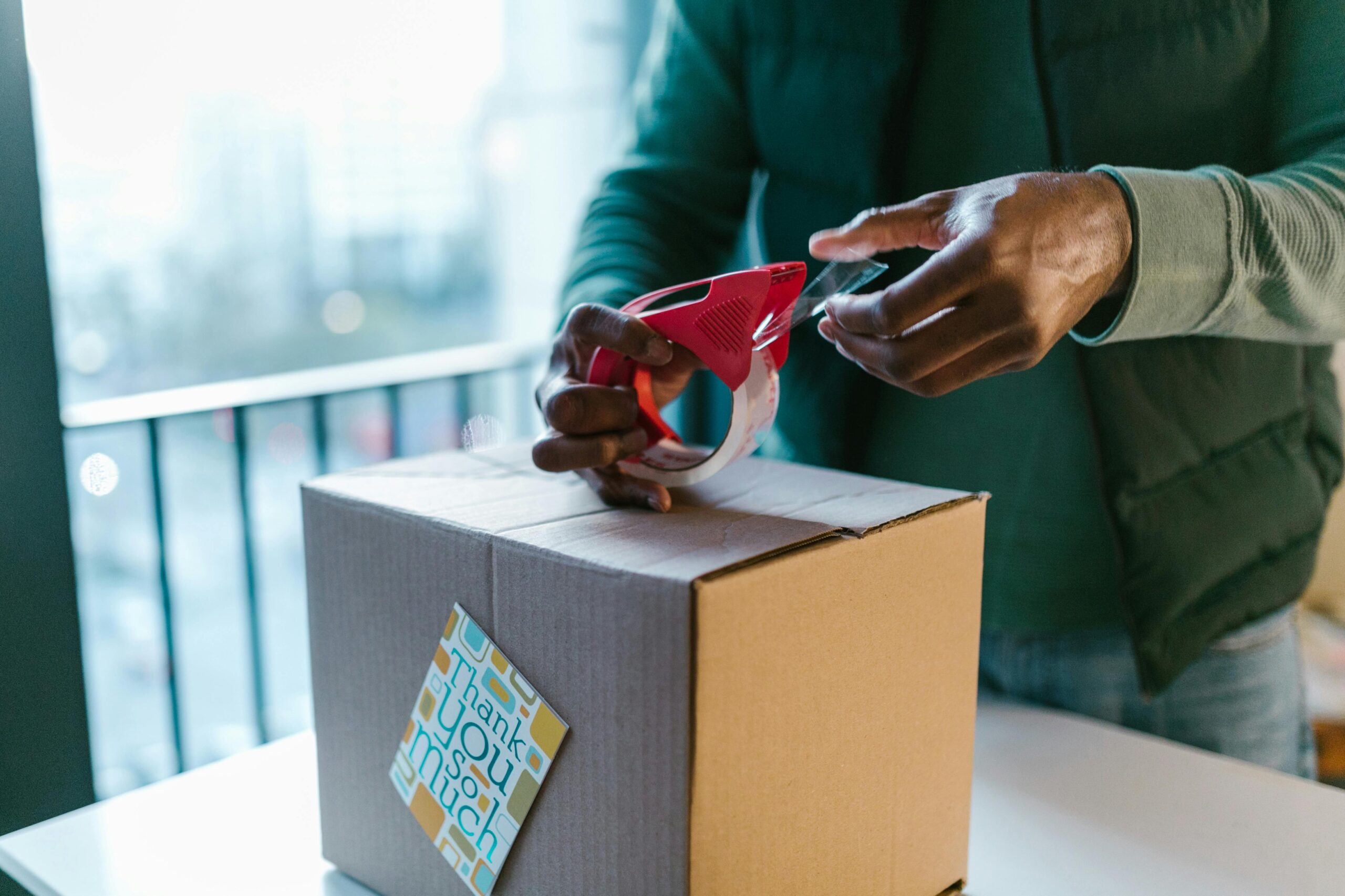 Close-up of man sealing a cardboard box with a tape dispenser indoors.